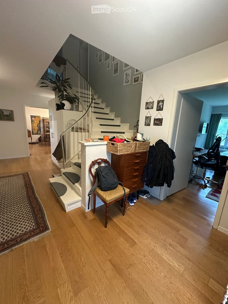 Spacious hallway with wooden flooring, staircase leading upstairs, coat rack, and a small seating area with a chair and backpack.
