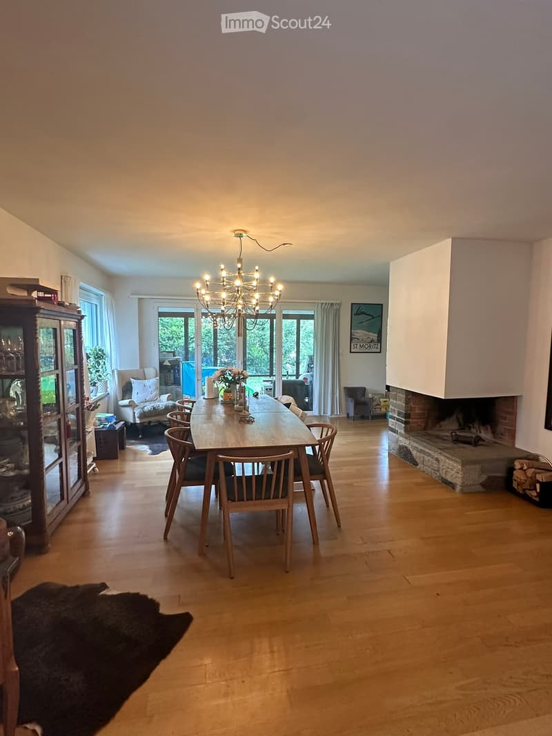 Bright dining room with wooden floor, large windows, chandelier, dining table with chairs, cabinet, and fireplace.