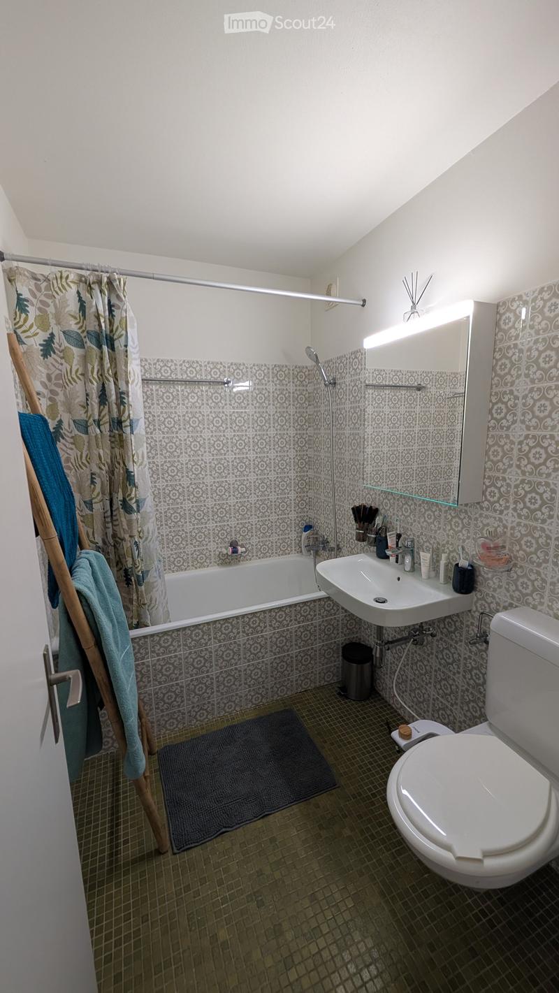 Bathroom with a bathtub, sink, toilet, mirror, and a shower curtain. The walls are tiled with a patterned design, and the floor is tiled with small square tiles. A towel hangs on a wooden rack, and a rug is placed in front of the bathtub.