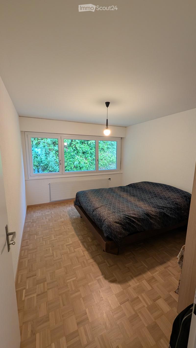Bedroom with wooden herringbone flooring, large window with three panes, bed with dark bedding, radiator below window, and a hanging light fixture.
