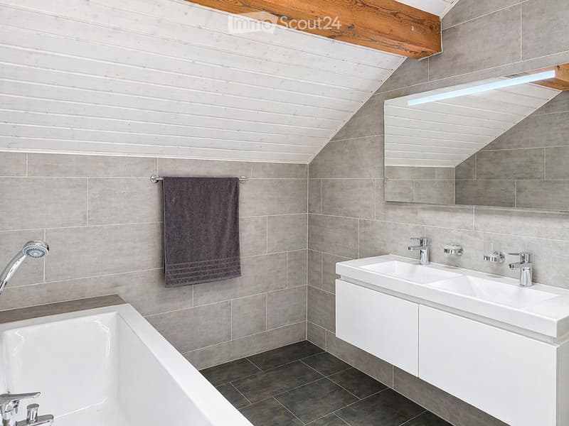 Bathroom with gray tiles, white bathtub, double sink, mirror, and wooden ceiling.