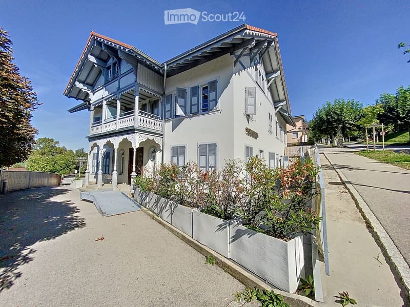 A two-story house with a red roof, white walls, and blue shutters. The house has a balcony on the second floor and a driveway in front. The house is surrounded by a garden with plants and trees.