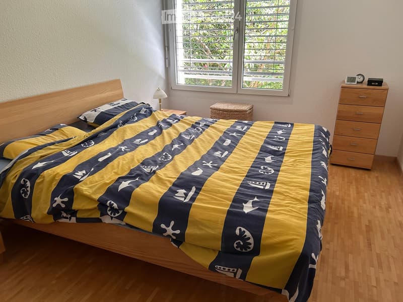 Cozy bedroom with wooden flooring, a single bed with a blue and yellow patterned bedspread, a wooden nightstand with a lamp, a wicker basket, and a wooden drawer unit with a clock on top.