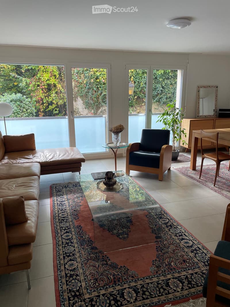 Modern living room with large glass doors leading to a garden, featuring a leather sofa, glass coffee table, armchair, dining table, and decorative vase.