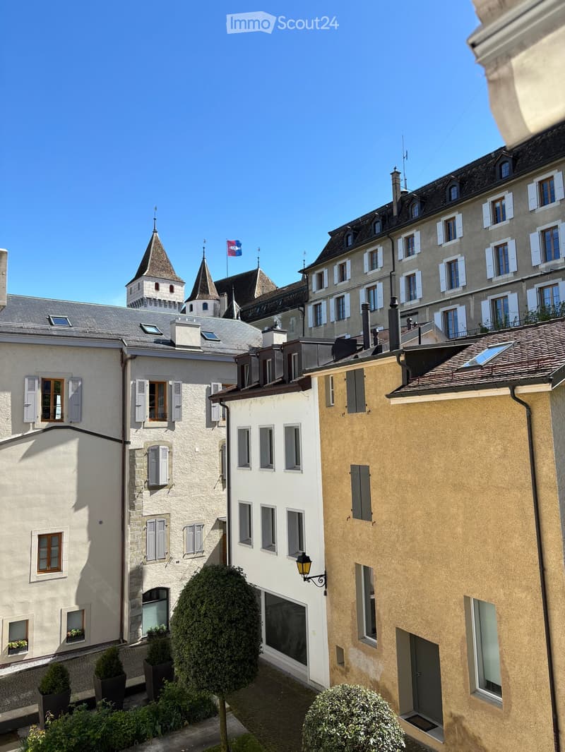 A picturesque European town scene with multi-story buildings, traditional architecture, and a clear blue sky. The buildings feature white and beige facades, shuttered windows, and ornate rooftops. A small garden area with trimmed bushes and potted plants
