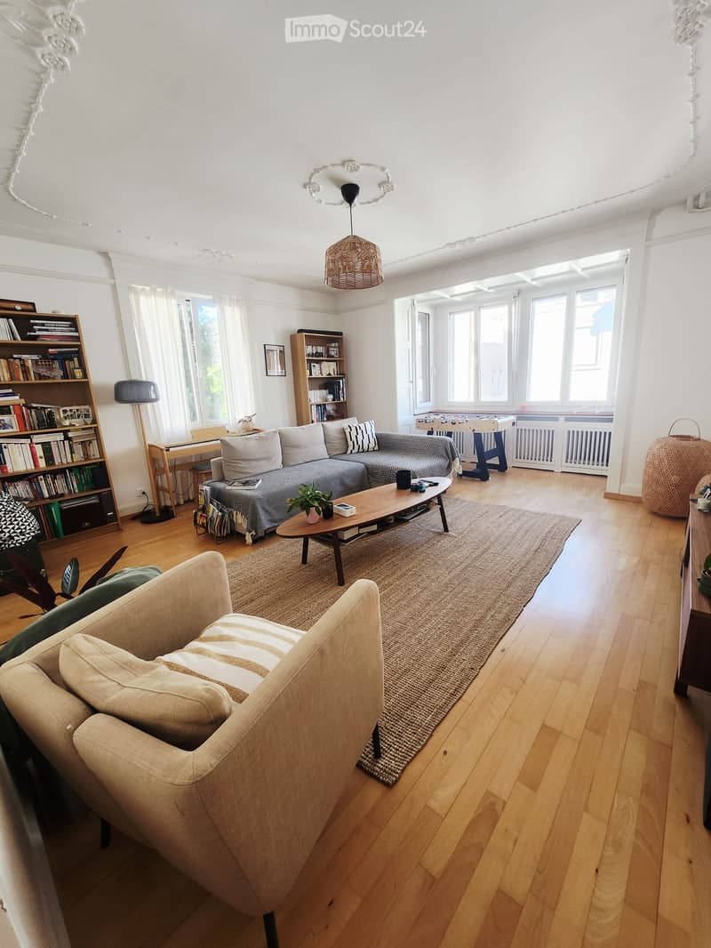 Bright living room with wooden flooring, large windows, a grey sectional sofa, a wooden coffee table, an armchair, bookshelves, and a football table in the background.