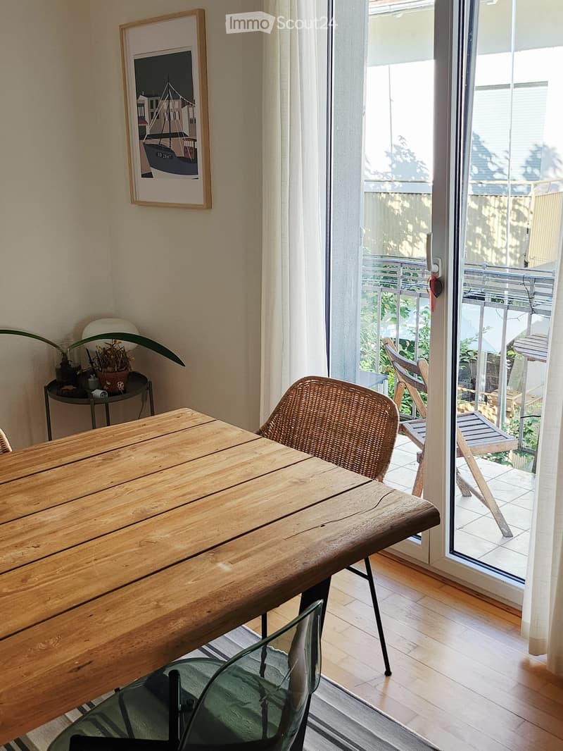 Bright room with wooden floor, large sliding glass door leading to a balcony, wooden table, wicker chair, glass chair, small plant, and picture frame on wall.