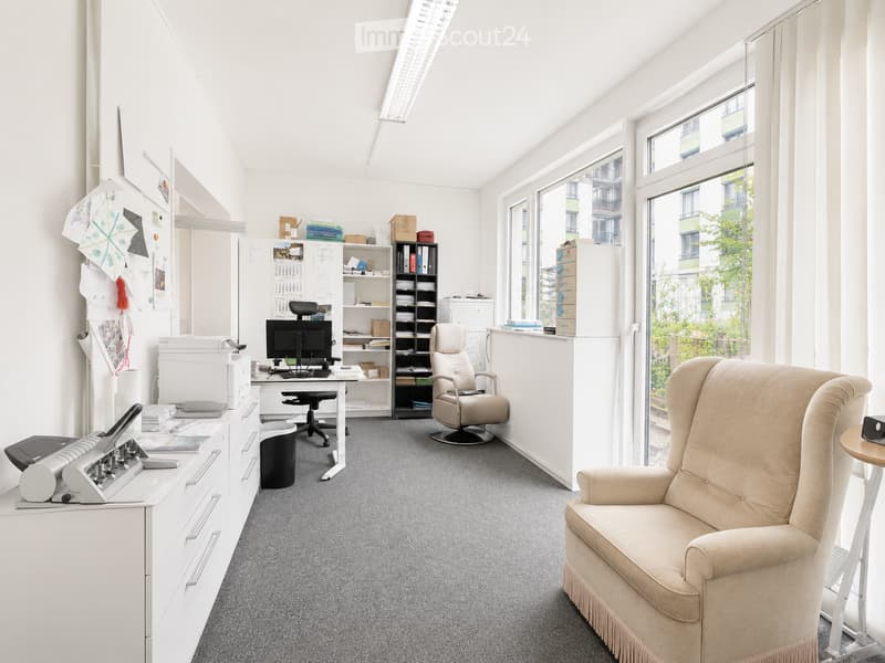 Bright office space with large windows, grey carpet, white walls, and modern furniture including a desk, chair, sofa, and printer.