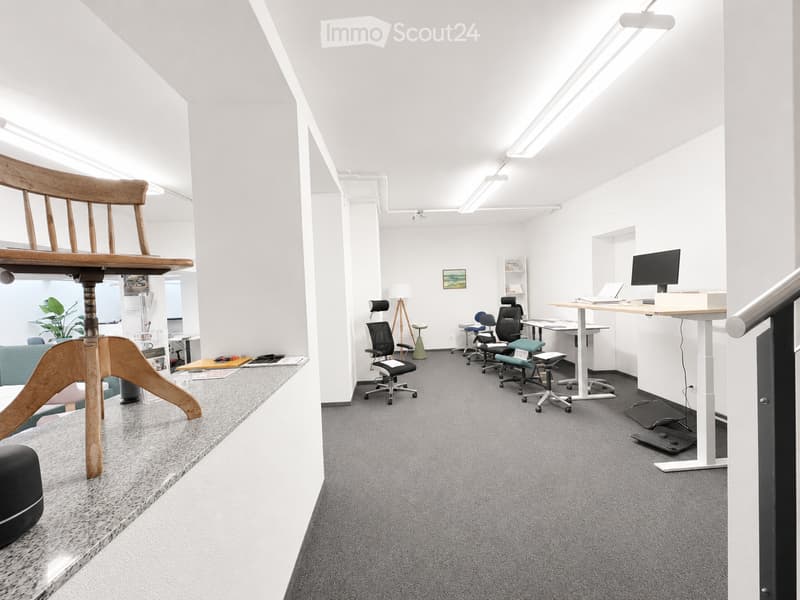 Modern office space with white walls, grey carpet, and various furniture including chairs, tables, and a laptop on a desk.