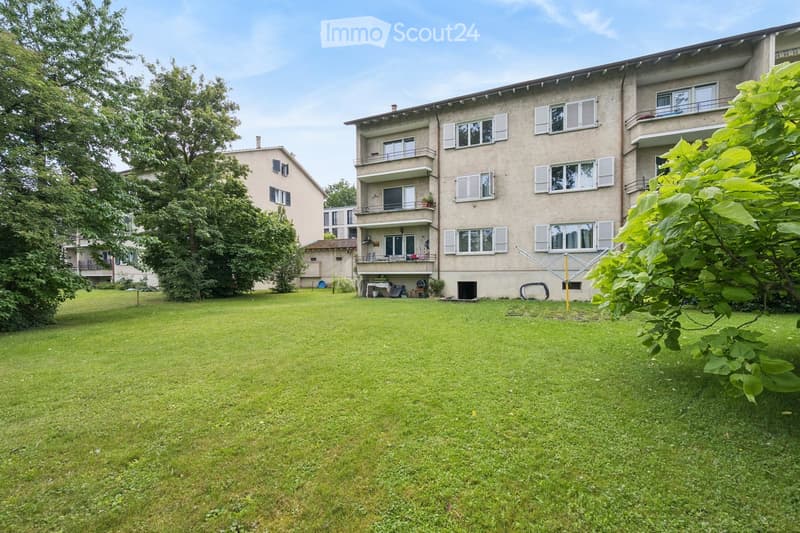 A well-maintained lawn in front of a multi-story residential building with balconies and windows. The building has a light-colored facade and is surrounded by trees and greenery.