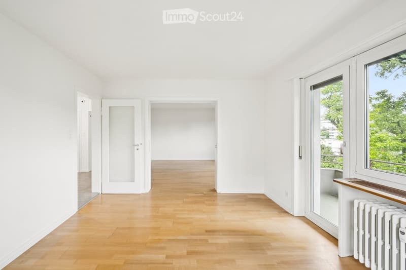 Bright, empty room with white walls, wooden flooring, and large windows offering a view of greenery. The space includes a radiator and a small wooden shelf near the window.