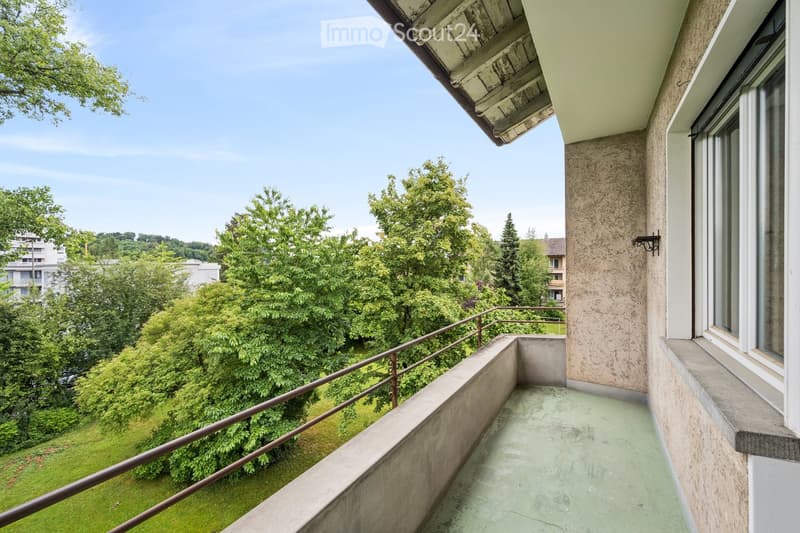 Balcony with metal railing, overlooking a green lawn and trees, with a building visible in the distance.