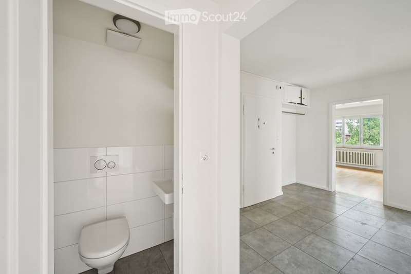 Modern bathroom with white tiles, toilet, sink, and built-in storage. Adjacent room visible with tiled floor and large windows.