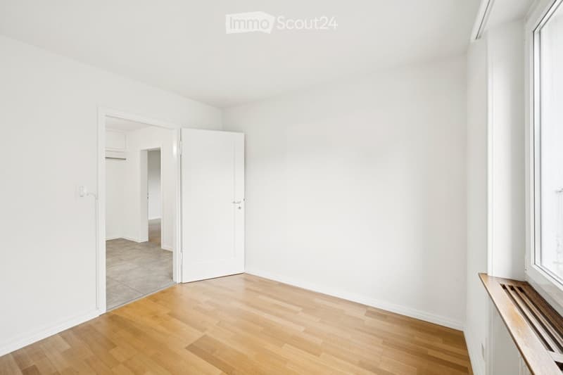 Empty room with white walls, wooden flooring, and a large window. A door leads to another room with tiled flooring.