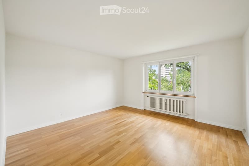 Empty room with light wooden flooring, white walls, and a large window with a radiator below it.