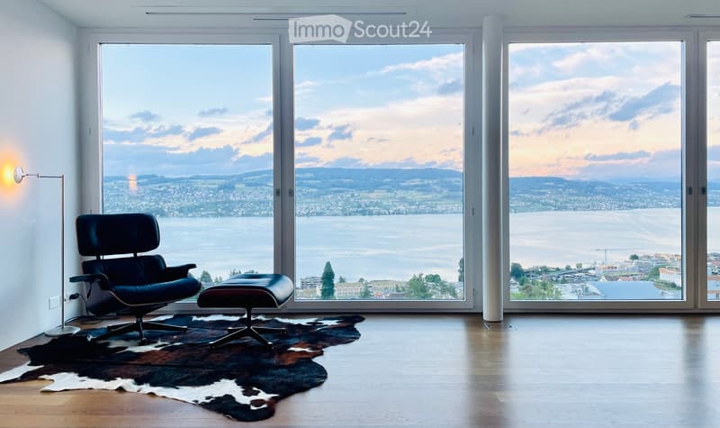 Furnished Room. Modern living room with large windows offering a scenic view, featuring a black leather chair, matching ottoman, floor lamp, and a cowhide rug on a wooden floor.