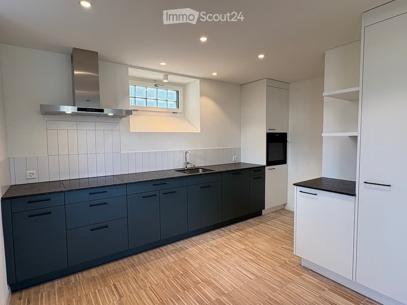 Modern kitchen with dark grey cabinets, white walls, and a wooden floor. Features include a sink, faucet, oven, and range hood.