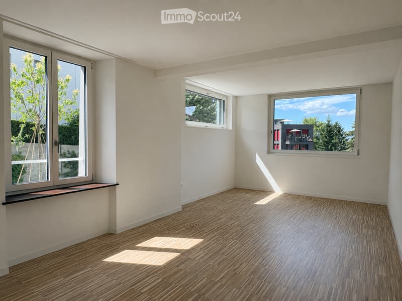 Empty room with light-colored walls, wooden flooring, and multiple windows offering views of greenery and a neighboring building.