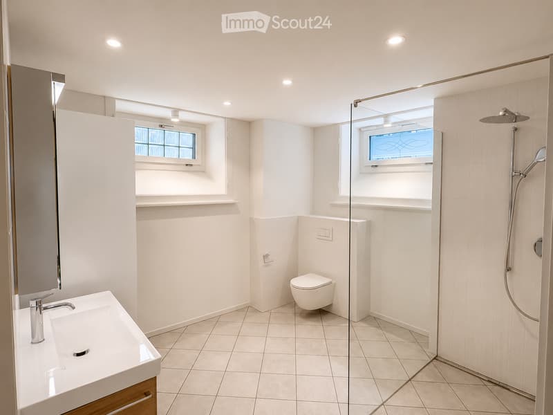Modern bathroom with white tiles, a glass-enclosed shower, a toilet, a sink with a chrome faucet, and a large mirror.