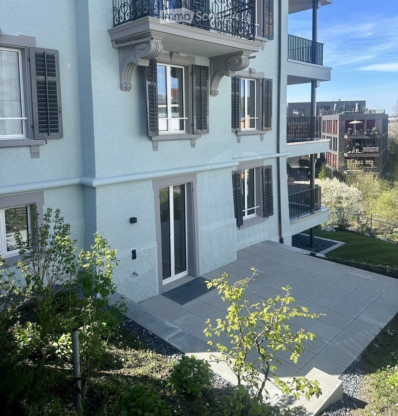 Modern multi-story building with light blue exterior, large windows, decorative shutters, and balconies. The ground floor features a tiled patio area with small plants and shrubs.
