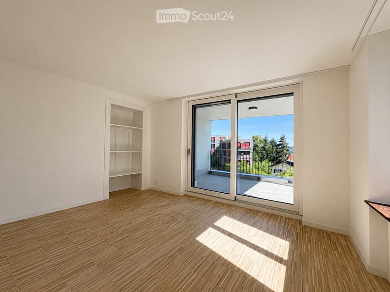 Empty room with wooden flooring, white walls, built-in shelving unit, and large sliding glass doors leading to a balcony with a view of trees and buildings.