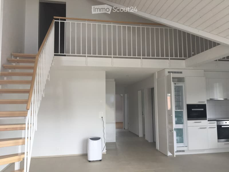 Open-plan living space with a staircase leading to an upper level, a modern kitchen area with white cabinets and stainless steel appliances, and a portable air conditioner on the floor.