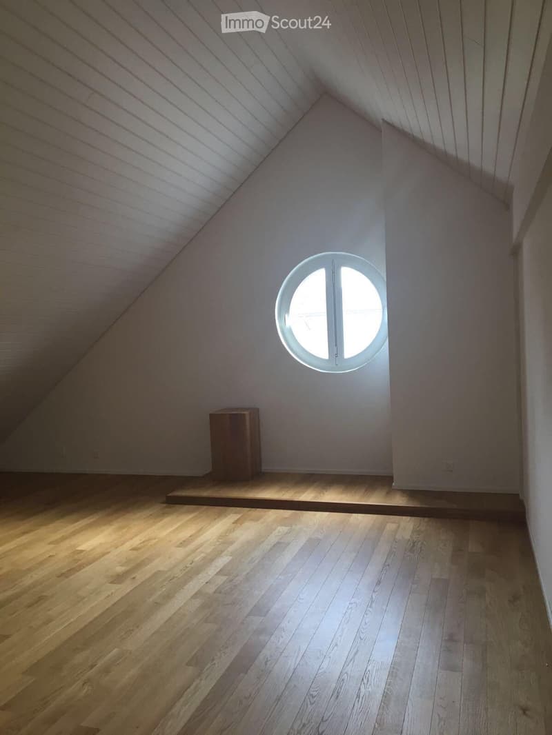 Empty attic space with wooden flooring, slanted wooden ceiling, and a round skylight window. A small wooden platform is visible against the wall.