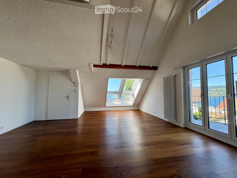 Bright, empty attic room with wooden flooring, white walls, and large windows offering a scenic view. The room features a sliding glass door leading to a balcony.