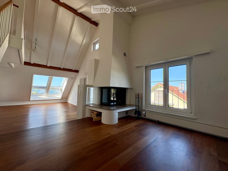 Bright, spacious attic room with wooden flooring, exposed wooden beams, large windows, and a built-in fireplace with a small storage compartment underneath.