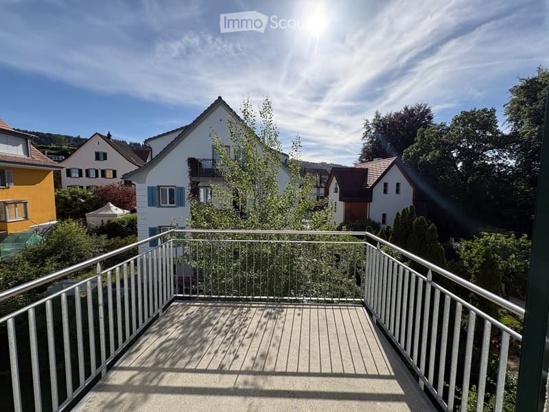 Balcony with metal railing, overlooking a residential area with houses, trees, and a clear blue sky.