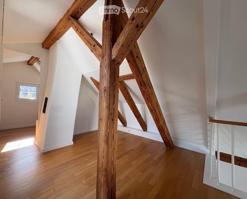 An attic space with exposed wooden beams, wooden flooring, and a small window letting in natural light.