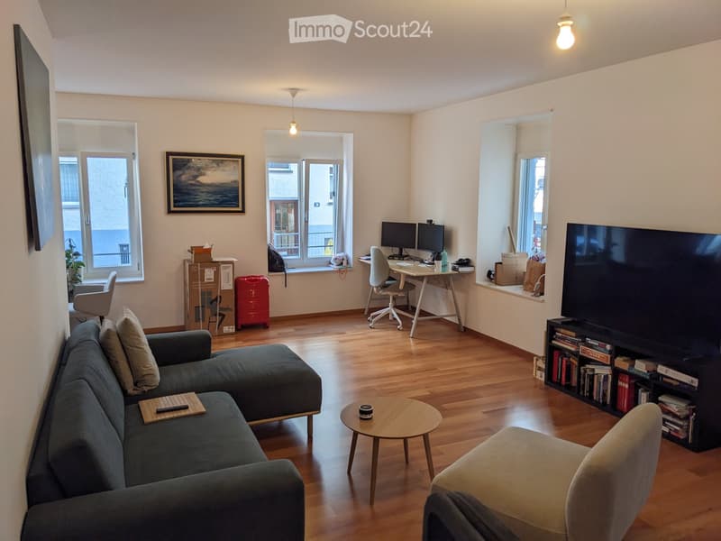 A modern living room with a grey sectional sofa, coffee table, armchair, desk with computer, and a large TV on a bookshelf. The room features wooden flooring and large windows with views.