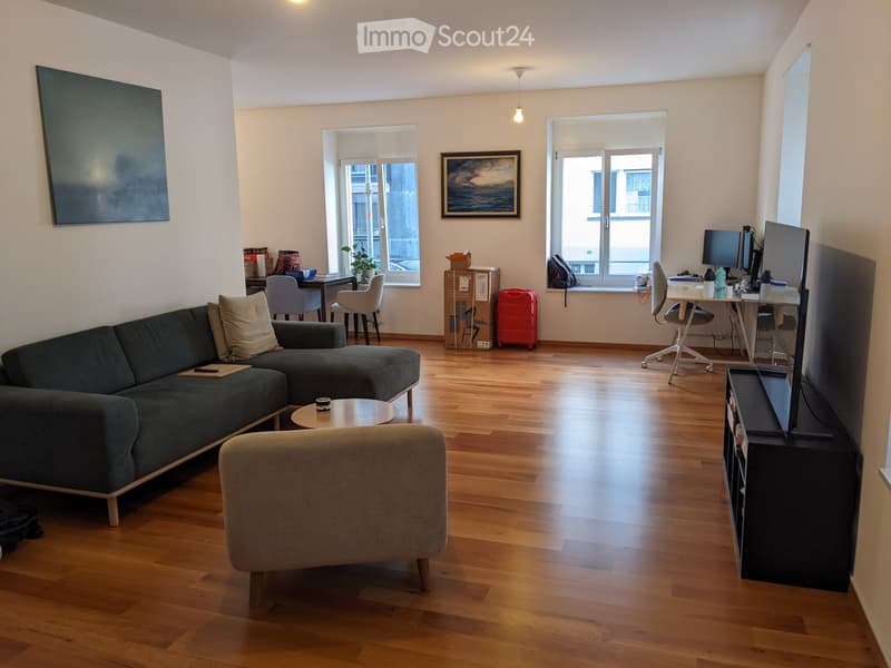 A modern living room with a dark green sectional sofa, a round coffee table, a grey armchair, a TV stand with a screen, a desk with a laptop, and a small table with a plant. The room has wooden flooring and large windows with blinds.