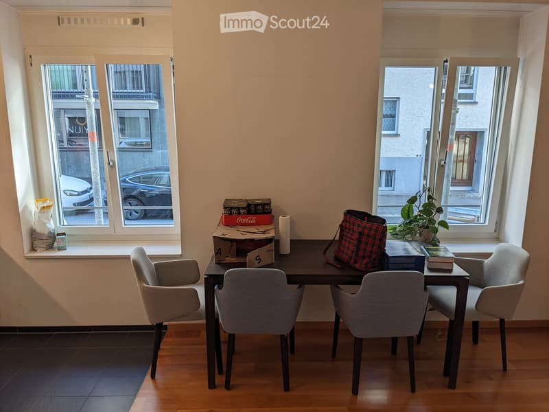 A small, furnished room with a wooden floor, a black dining table surrounded by four grey chairs, a handbag, a potted plant, and some cardboard boxes on the table. The room has large windows with a view of the street outside.