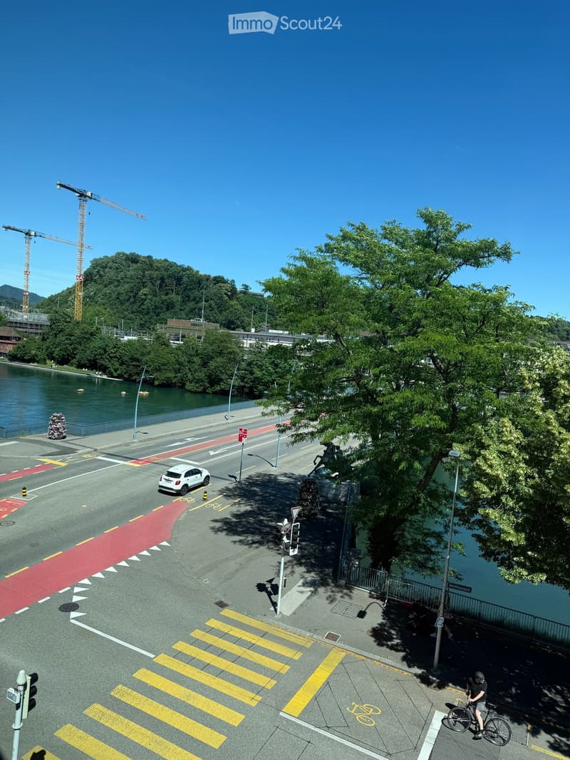 Aerial view of a riverside area with a road, pedestrian crossing, bicycle lane, and trees. A white car is visible on the road, and a person is riding a bicycle near the water.