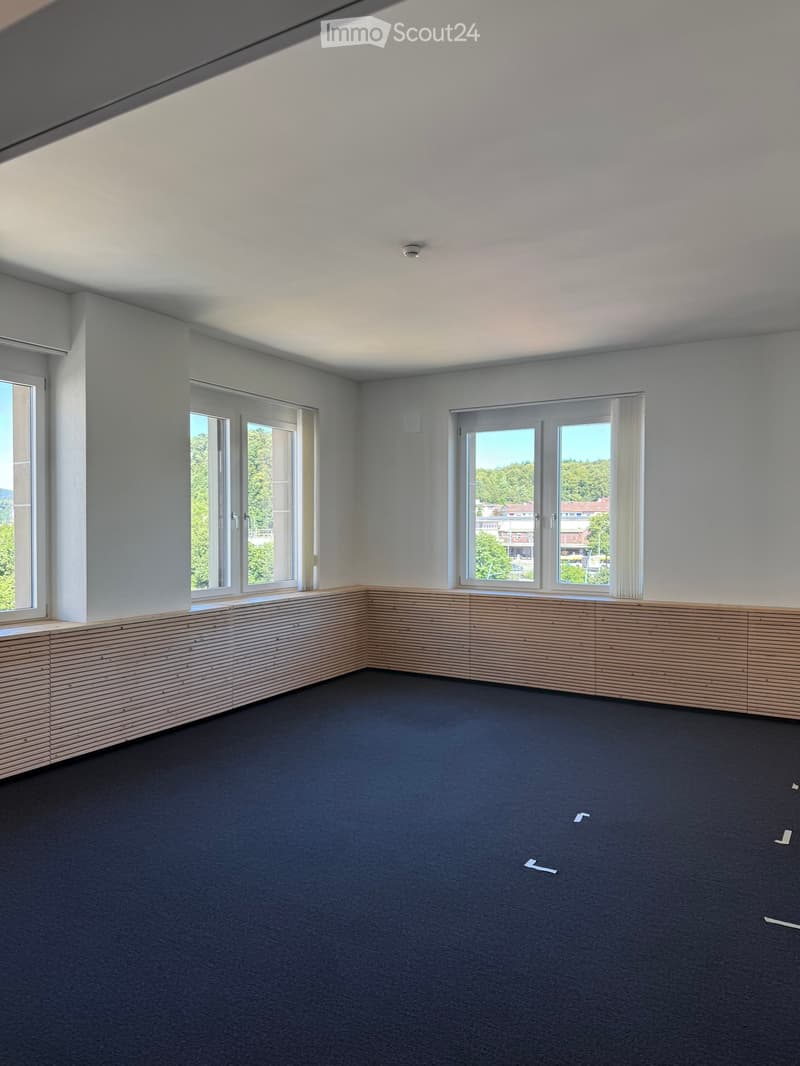 Empty room with grey walls, dark carpet, large windows, and a wooden bench along the wall.
