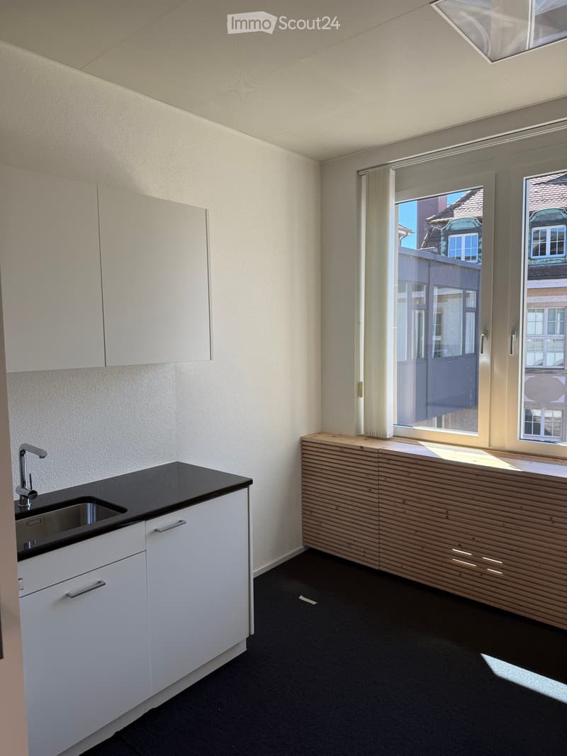 Modern kitchen with white cabinets, black countertop, and a wooden breakfast bar. Features a sink with a modern faucet and large windows offering a view of the street.