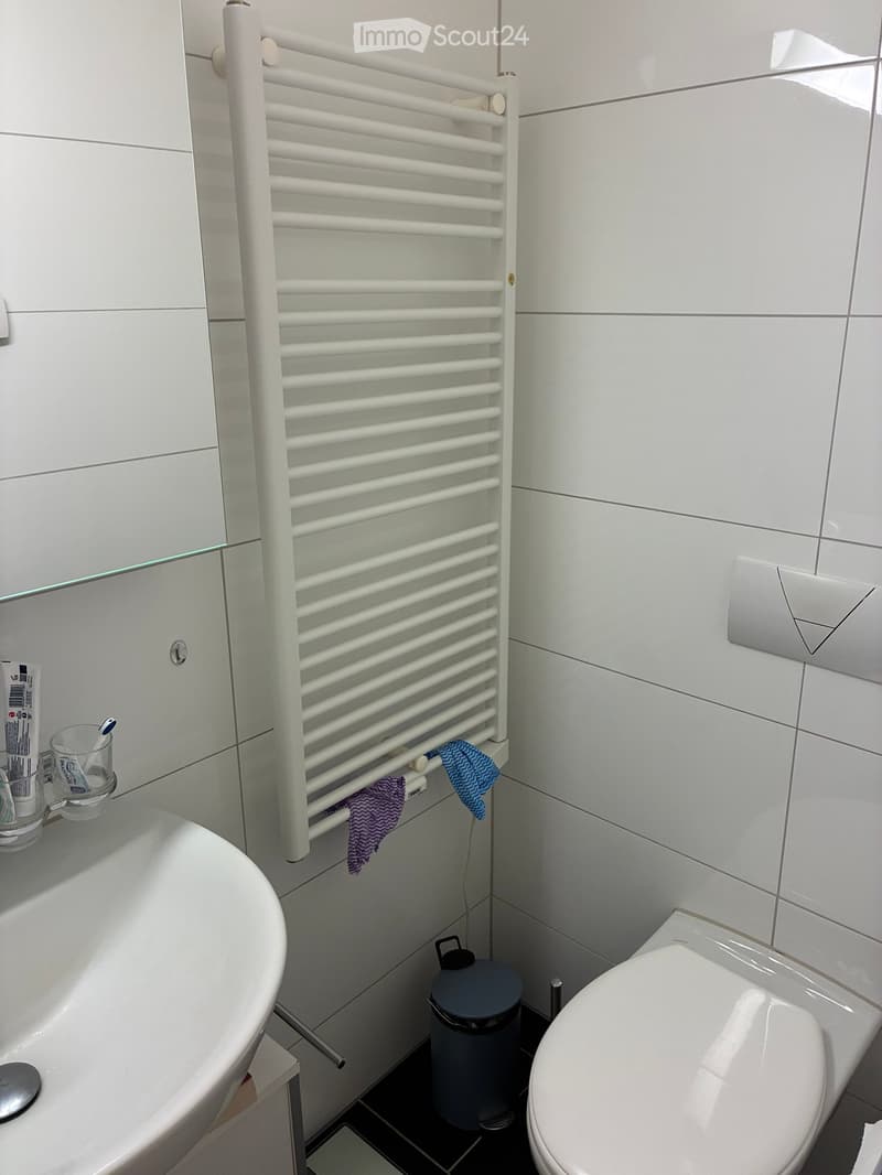 Modern bathroom with a white bathtub, toilet, toothbrush holder, toothpaste, and a towel rack with towels. The walls are tiled in white, and there is a small trash bin near the floor.