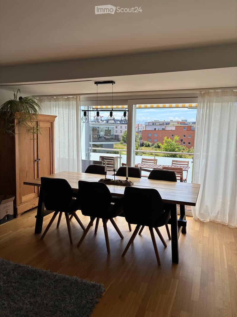 Dining room with wooden floor, large dining table, six chairs, pendant lights, and a view of the balcony and cityscape.