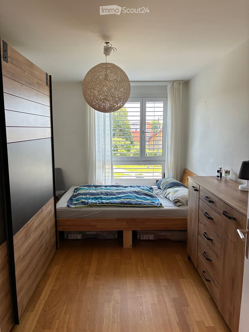 Cozy bedroom with wooden flooring, a single bed with a blue and white striped bedspread, a nightstand with a lamp, and a drawer unit. The room features large windows with white curtains and a modern pendant light.