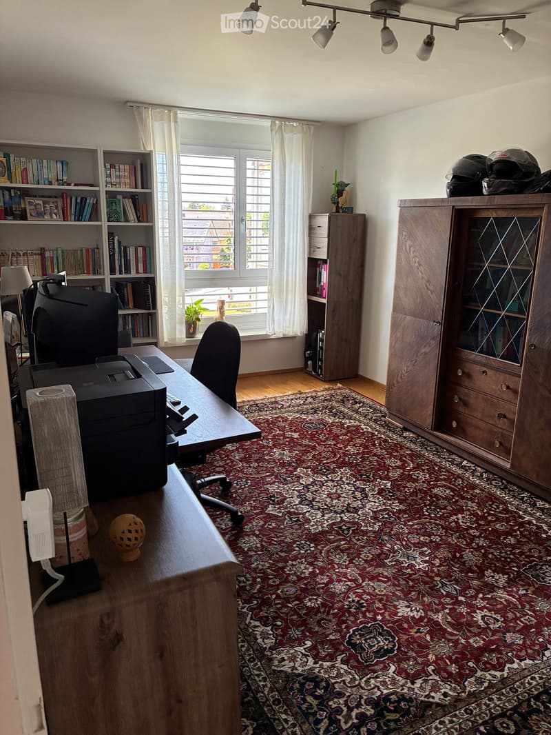 Well-lit study or home office with wooden flooring, large window with white curtains, patterned rug, desk with chair, bookshelf, cabinet, printer, and decorative items.