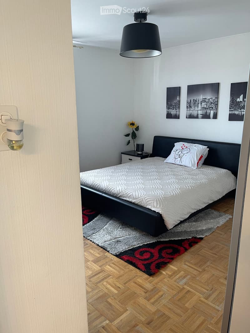 Modern bedroom with a double bed, nightstand, lamp, and artwork on the wall. The room features wooden flooring and a cozy rug.