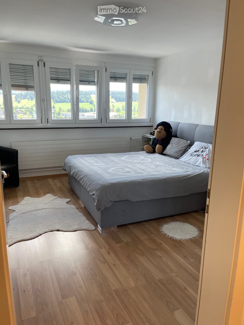 Modern bedroom with wooden flooring, large windows offering a scenic view, a grey bed with white bedding, a stuffed monkey toy, and a white fur rug on the floor.