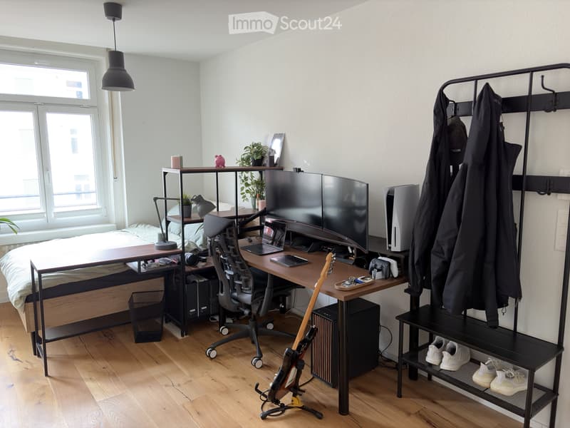 Modern, multi-functional room with a lofted sleeping area, a desk, and storage shelves. Features a sleek black chair, a wooden table, a guitar, and a modern lamp on the floor.