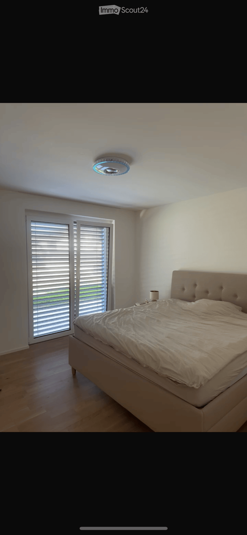 Modern bedroom with a large bed, nightstand, and lamp, featuring large windows with blinds and a sleek ceiling light fixture.
