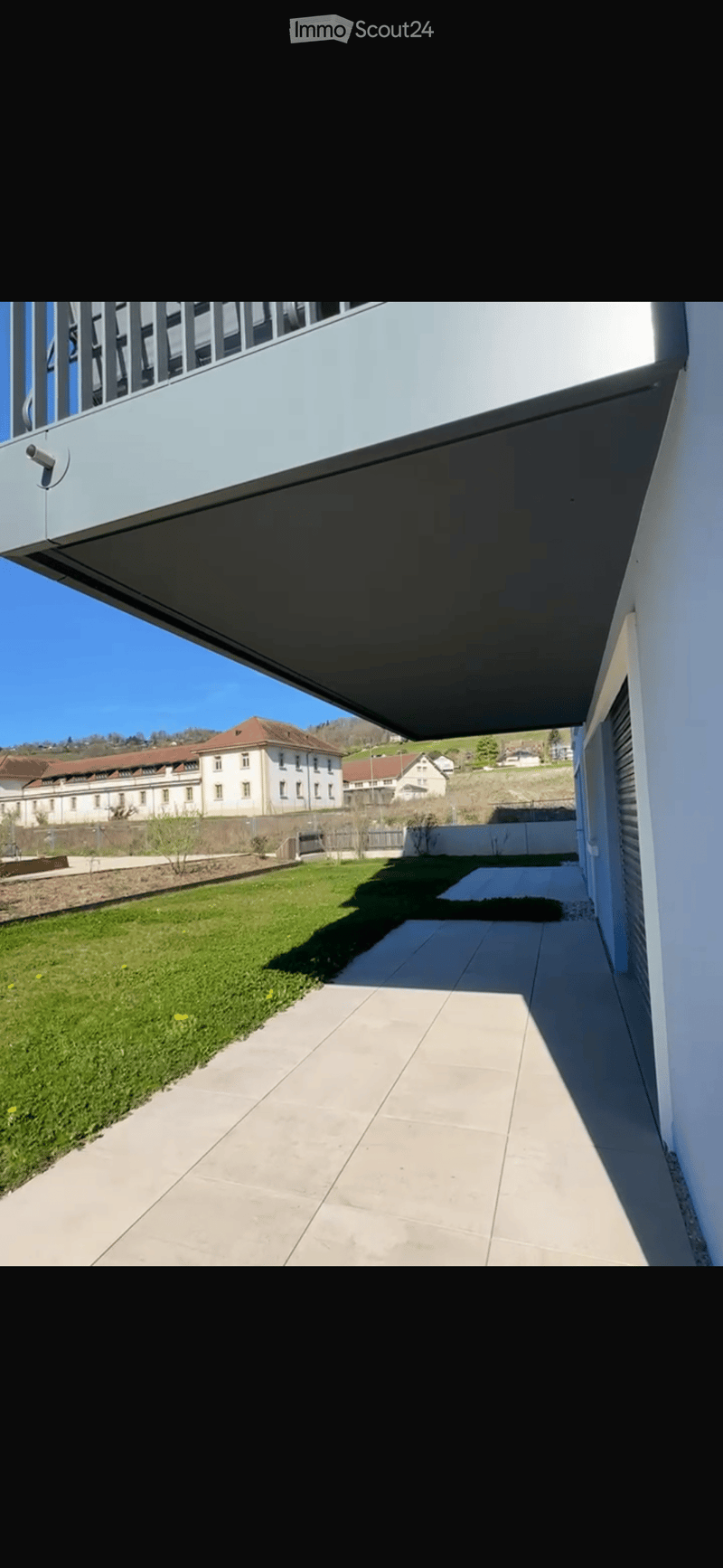 A modern outdoor area with a paved walkway, grassy lawn, and a view of a distant building complex.