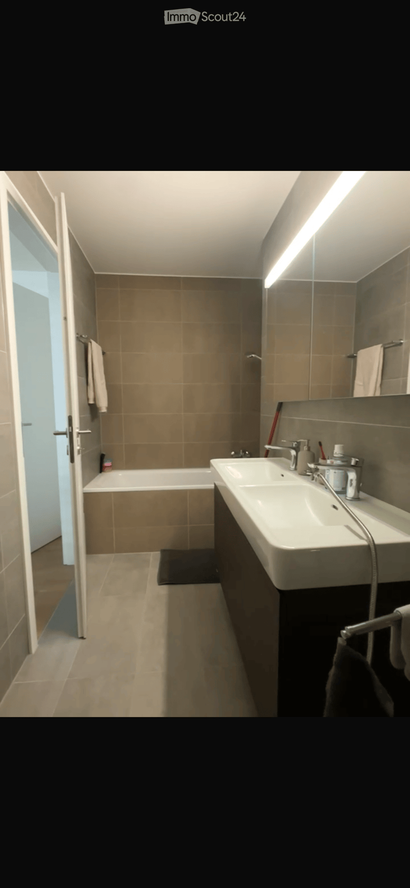 Modern bathroom with a bathtub, double sink, large mirror, and towel rack. The space features beige tiles and a sleek design.