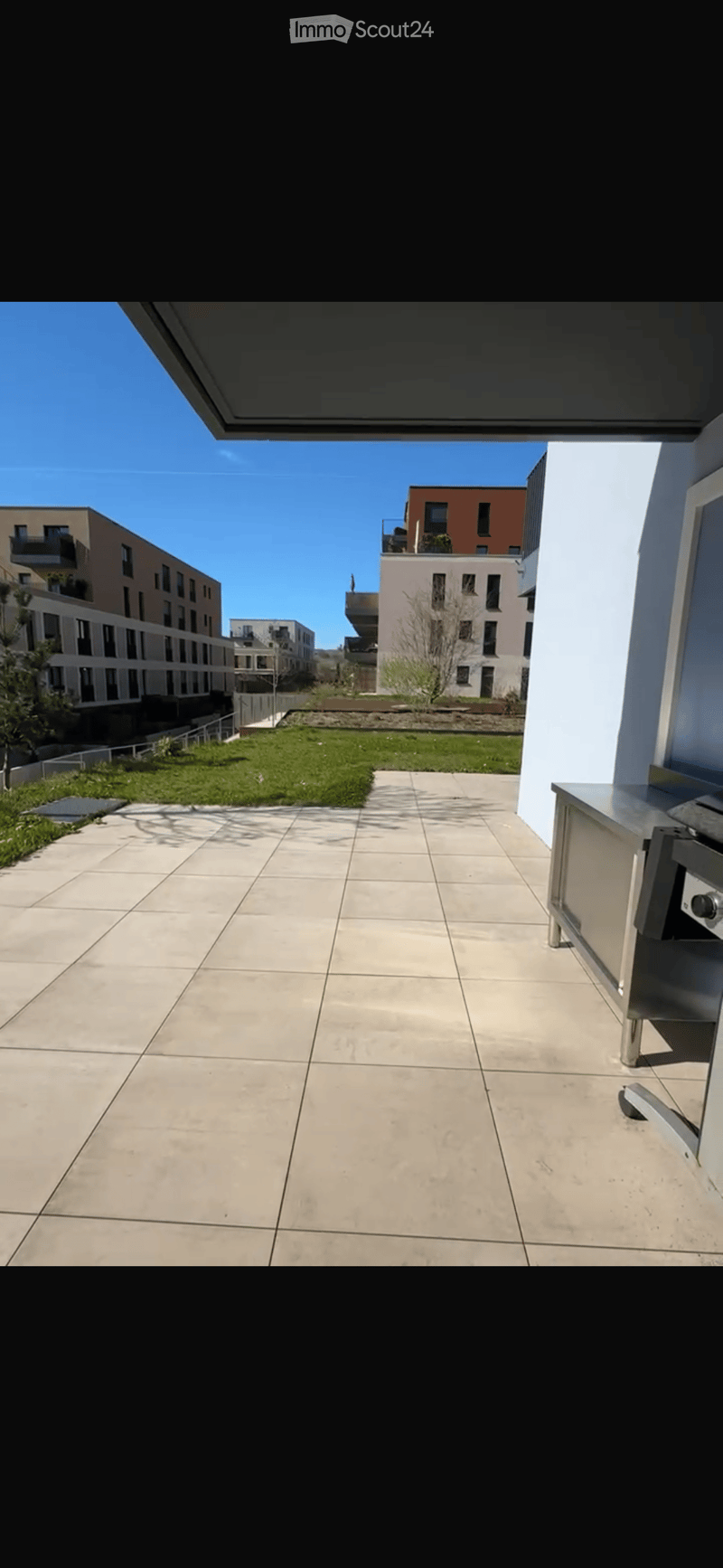Spacious balcony with tiled floor, modern stainless steel railing, and a view of a landscaped area with grass and modern apartment buildings.