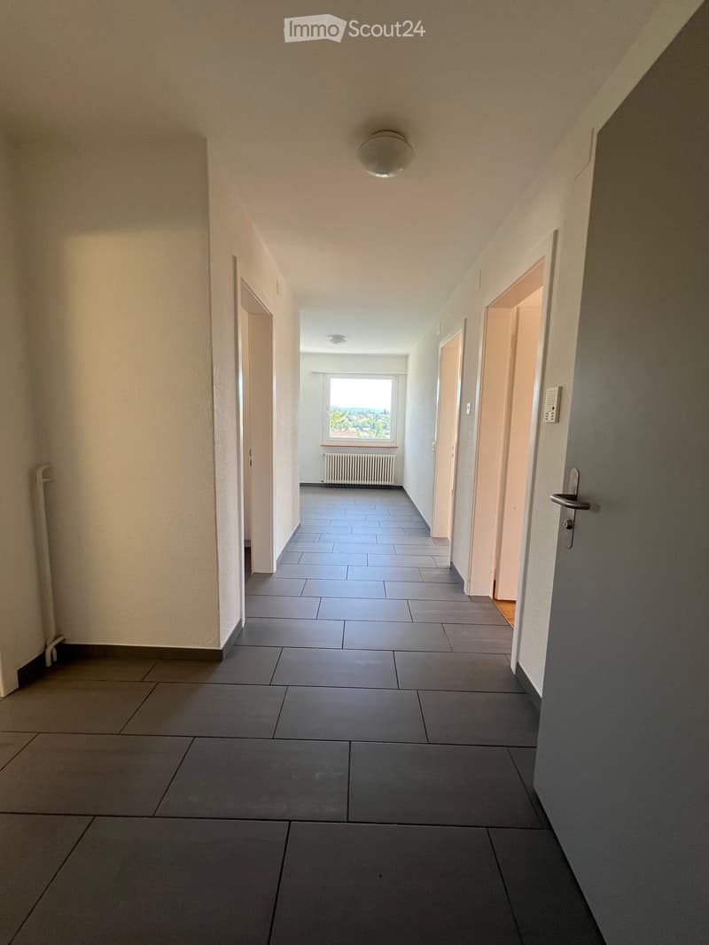 A long, narrow hallway with grey tiled flooring, light-colored walls, and several doors on both sides. A window at the far end provides natural light, and a radiator is visible beneath it.
