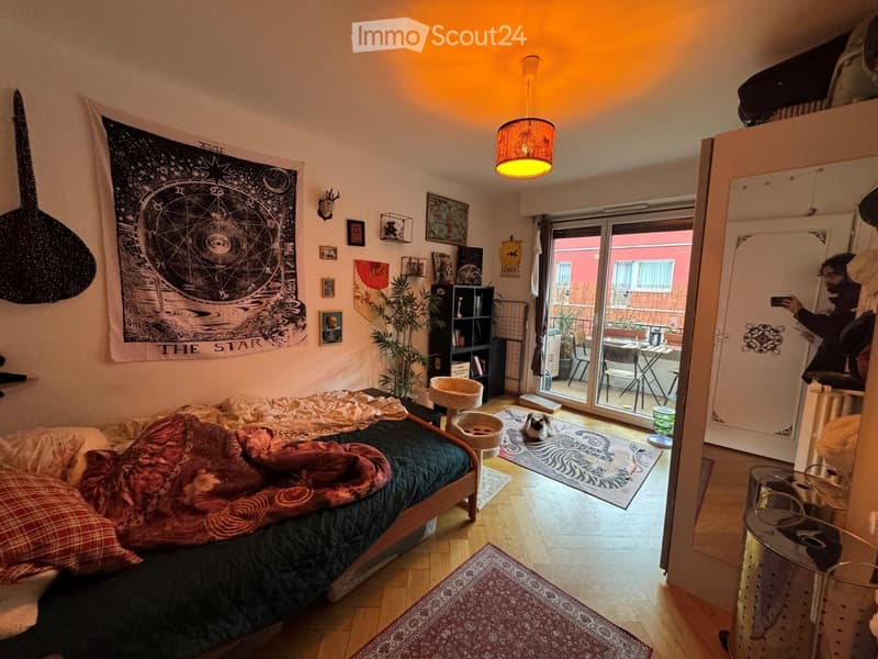 Cozy bedroom with a wooden bed, colorful blankets, a cat tree, a small cat, a bookshelf, a hanging lamp, and a sliding glass door leading to a small outdoor area with a table and chairs.